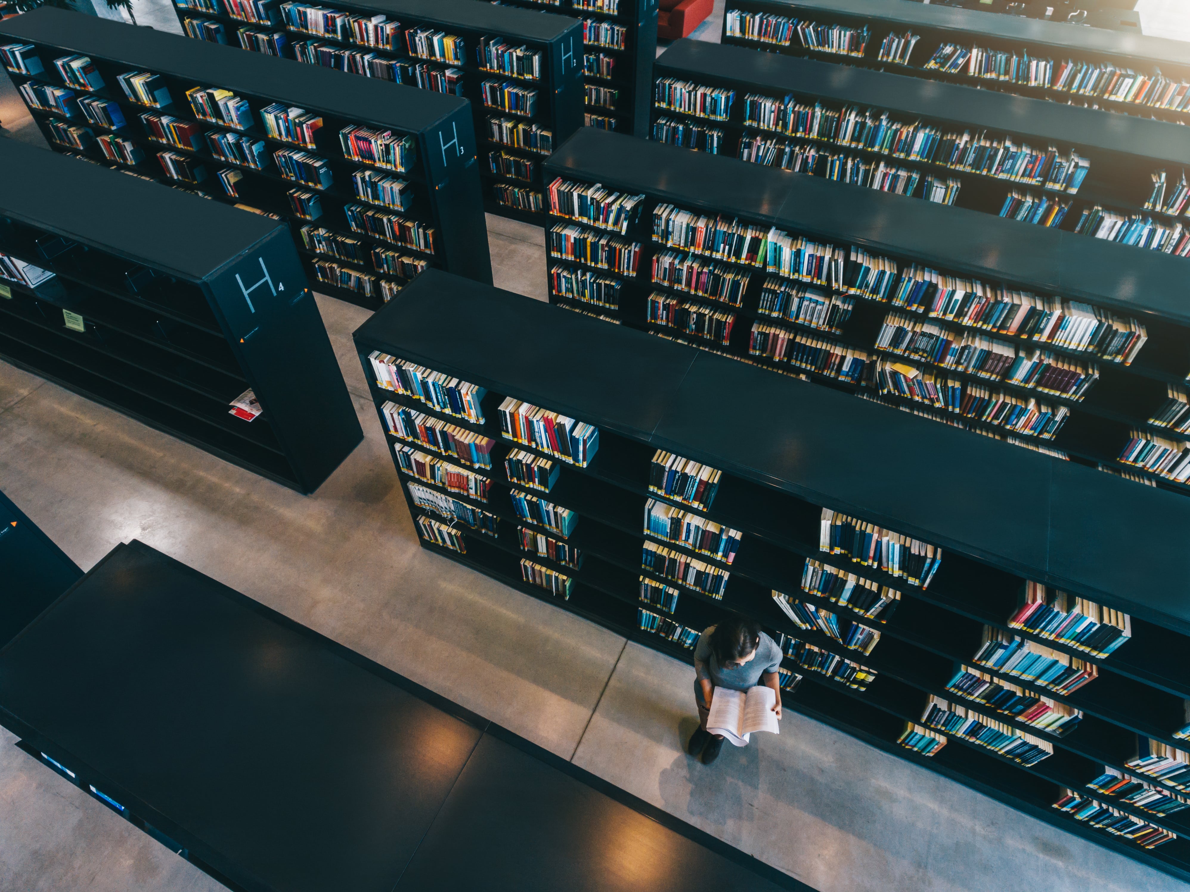 female-student-at-library-bookshelf-reading-book-PRN3AZU-min